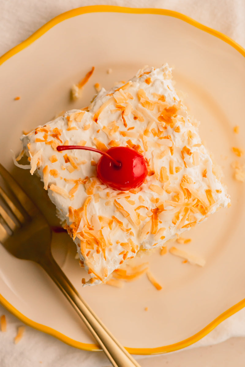 Slice of cake with coconut and a cherry on a beige plate with a gold fork.